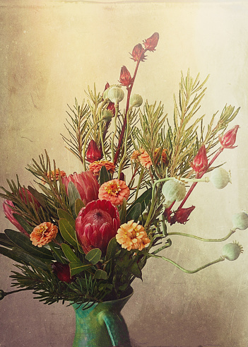 My original vertical closeup photo of a green pottery jug filled with a bright bouquet of orange Zinnias, green Poppy seedpods, red Rosella plant seed heads, red Protea flowers and branches of green Banksia leaves, has been transformed using the Mextures App to give a retro, vintage, faded old world look to the image. Textured, beige and cream toned background.