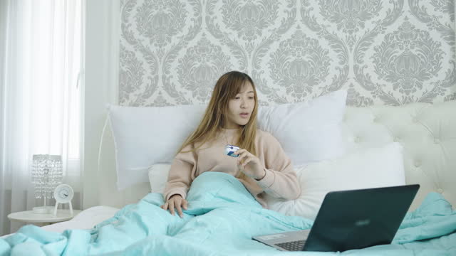 Young Asian woman sitting in bed using her notebook as she uses fever scanner and oximeter