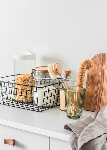 Kitchen organization interior - cans of pasta, flour in a metal basket, brushes for washing dishes, cutting boards, napkin on a white table in a bright room