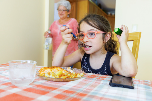 Funny little girl in eyeglasses eating dinner in the kitchen, holding cucumber in her hand.