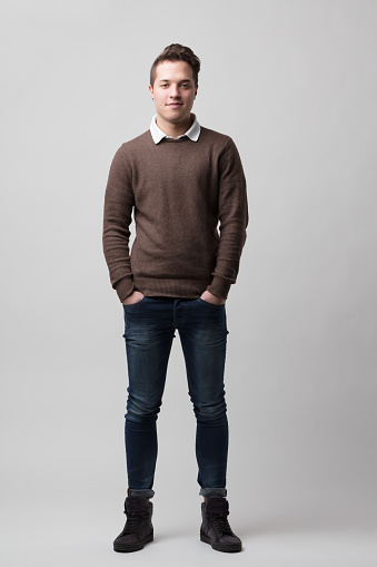 Portrait of relaxed young man with short hair and topknot fashionable in the 2020s. Wearing ankle-high skinny jeans and dark gray ankle boots, the collar of a light-colored shirt pokes out from the br