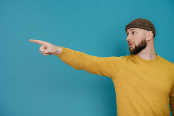 istock Go back! Young handsome caucasian beardy man in hat and yellow sweater pointing by index finger away against turquoise studio background with copy space.  Confident guy shows motion direction. 1473837741