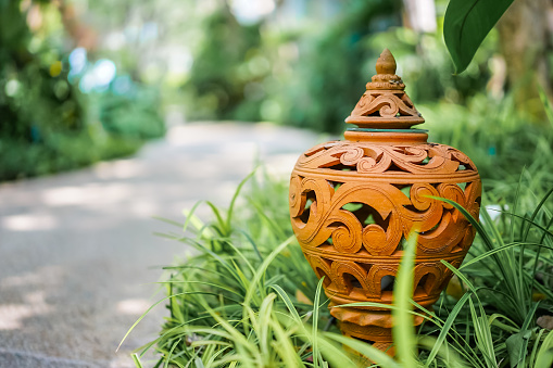 The close up photo of the outdoor handmade carving ceramic clay lamp in the exquisite Thai style pattern settled on the grass of the garden near the passage.
