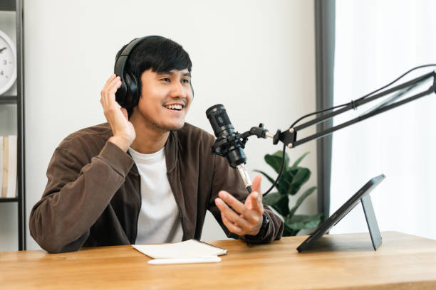 istock Young asian man host streaming podcast with condenser microphone work on tablet at small broadcast home studio. Content creator blogger recording voice over radio interview guest conversation 1473840142