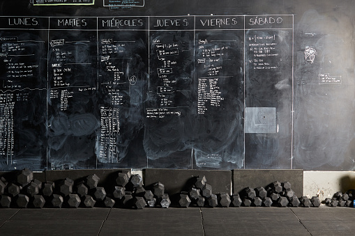 Stock photo of a workout plan written with chalk on a blackboard with a lot of weights underneath it. There are nobody on the picture.
