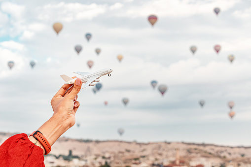 A hand holds a toy airplane with Cappadocia's hot air balloons in the background, representing air transport and travel concept.