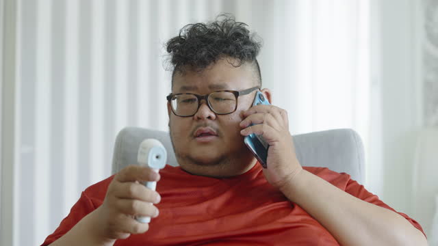 Overweight Asian man coughing uses thermometer measuring his temperature then quickly calls to ask for sick leave