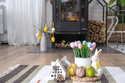 Key to house near fireplace stove with fire and firewood. Cozy home hearth with easter decor, colorful eggs in a basket and bunny. Building, moving, mortgage, rent and purchase real estate, insurance