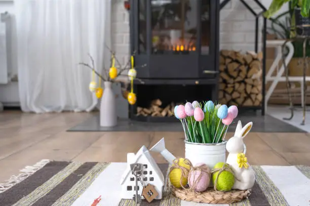 Photo of Key to house near fireplace stove with fire and firewood. Cozy home hearth with easter decor, colorful eggs in a basket and bunny. Building, moving, mortgage, rent and purchase real estate, insurance