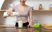 Smiling young woman pouring milk into a glass while sitting and having breakfast at the kitchen table.