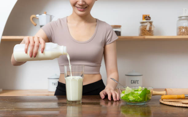 Smiling young woman pouring milk into a glass while sitting and having breakfast at the kitchen table. stock photo