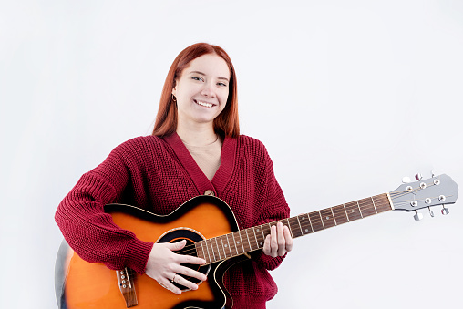 young caucasian red-haired woman in red sweater playing acoustic guitar isolated on white background. Guitar courses ad