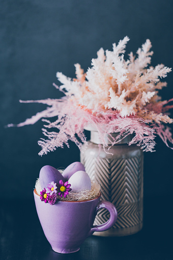 Dark low key photo of a rustic purple cup with daisies, feathers and purple Easter eggs in the foreground and a vase with dried pink and beige shrubs like pampas grass on the black background. Creative color editing with added grain. Very soft and selective focus. Part of a series.