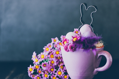 Dark low key of an Easter bunny cookie cutter in a cozy rustic purple cup with daisies, feathers and Easter eggs. Creative color editing with added grain. Very soft and selective focus. Part of a series.
