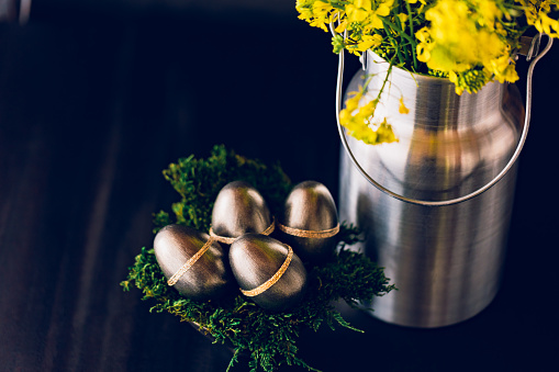 Dark low key still life of metallic painted Easter eggs and a silver colored milk pot as a vase with a oilseed rape bouquet. Copy space. Creative color editing with added grain. Very soft and selective focus. Part of a series.