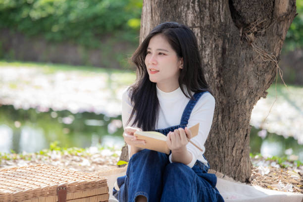 Beautiful young woman wearing denim overalls sitting in park reading book. picnic concept.