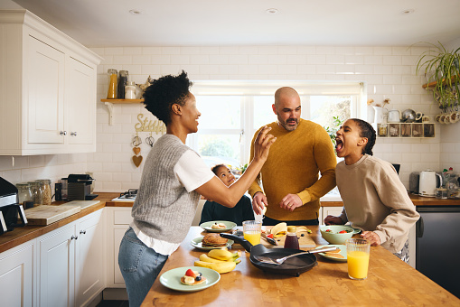 Mother aiming to throw food towards daughter's open mouth at family breakfast