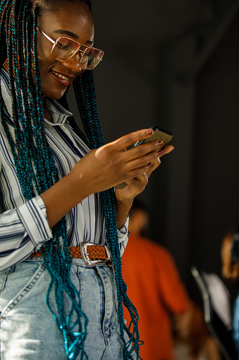 Close up shot of charming young Black businesswoman standing in a modern office space, taking a break from work and responding to a text message via smart phone.