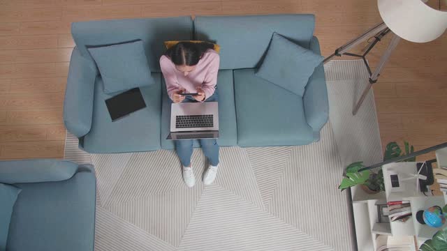 Top View Of A Woman Playing Game On Smartphone While Working With Laptop On Sofa At Home