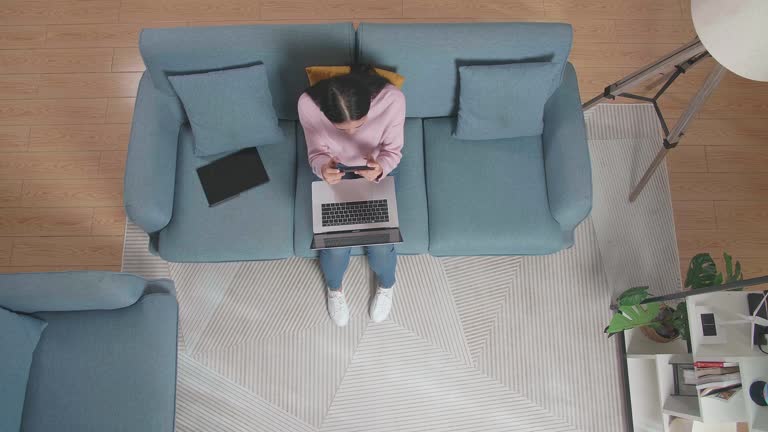 Top View Zoom Out Shot Of A Woman Playing Game On Smartphone While Working With Laptop On Sofa At Home