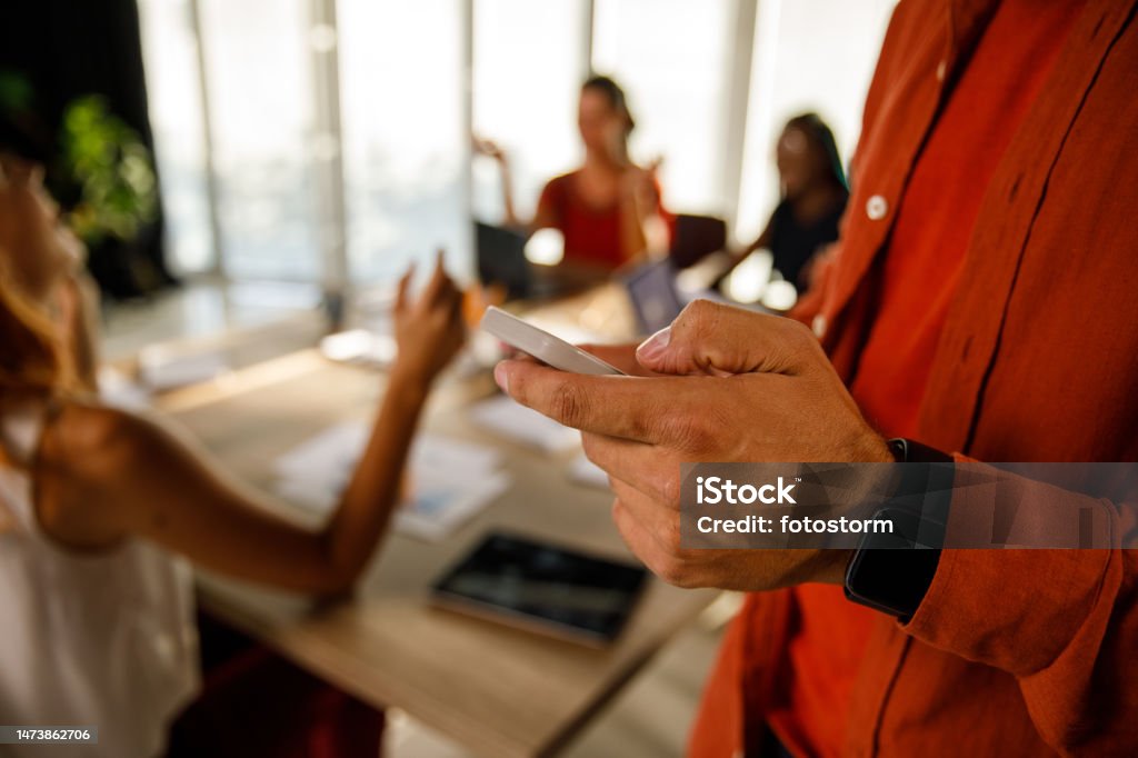 Businessman standing in a modern office, responding to a text message via smart phone Close up shot of unrecognizable businessman standing in a modern office, responding to a text message via smart phone. His colleagues having a meeting in the background. Smart Phone Stock Photo