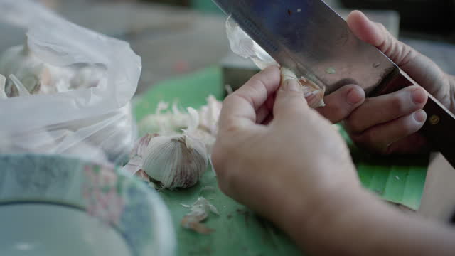 Senior woman hand peeling garlic with knife on wooden chopping board.