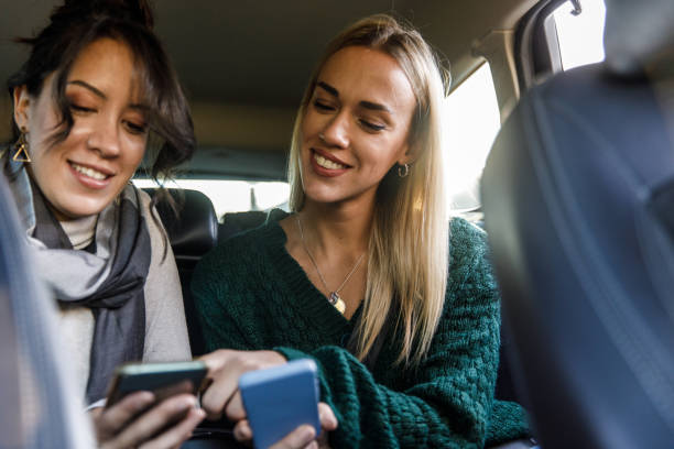 Two young women riding in the back seat of a taxi and showing each other memes and data on smart phones stock photo