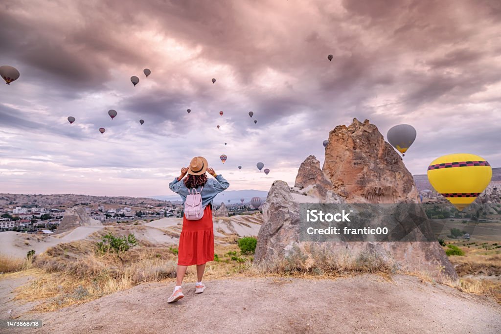Young woman stands in awe beneath the magical sight of hot air balloons soaring in the sky above the beautiful landscape of Cappadocia. She is dressed in a stylish dress and hat 30-34 Years Stock Photo
