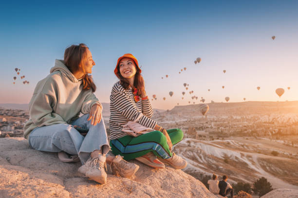 Girl friends sitting on a clifftop viewpoint and admiring view of majestic flying hot air balloons in Cappadocia Girl friends sitting on a clifftop viewpoint and admiring view of majestic flying hot air balloons in Cappadocia wow airlines stock pictures, royalty-free photos & images