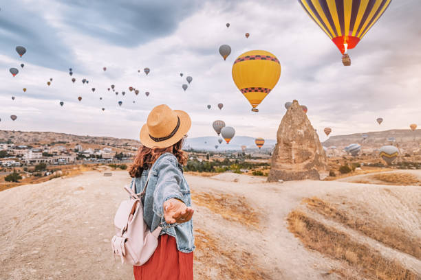 トルコのカッパドキアでの冒険に参加して、若い女性が自信を持ってフォローミーポーズをとり、熱気球を背景に舞い上がります。 - travelogue ストックフォトと画像