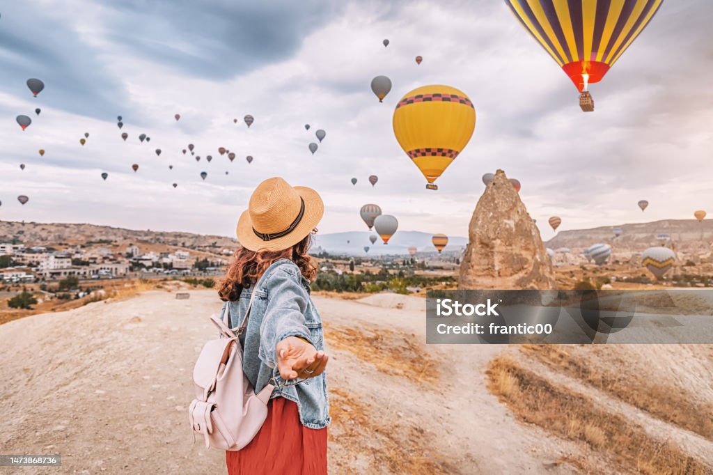 Join the adventure in Cappadocia, Turkey as a young woman strikes a confident follow me pose with hot air balloons soaring in the background. Cappadocia Stock Photo