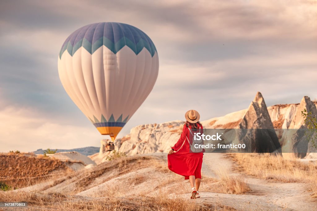 The air was alive with the sight of the balloons hovering in the sky above, and the girl below them in her red dress shining in the sunlight. She stood there, captivated. Cappadocia Stock Photo
