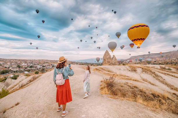 two girls friends capture the beauty of cappadocia, turkey, with their smartphones amid stunning rock formations and hot air balloons. - color image people air vehicle airplane imagens e fotografias de stock