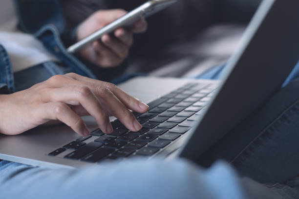 istock Young woman sitting on sofa using mobile phone and laptop computer at home, close up 1473874180