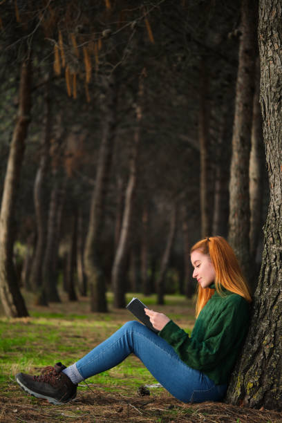 istock Redhead young woman reading a book sitting under tree in forest. 1473876668