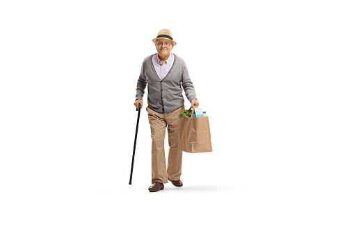 Full length portrait of an elderly man with a cane walking and carrying a paper grocery bag isolated on white background