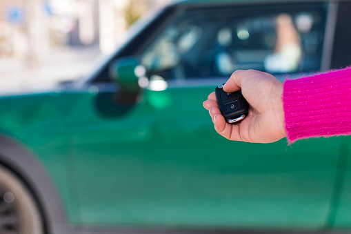Crop female in pink sweater pressing button on remote car key while opening green vehicle against blurred background