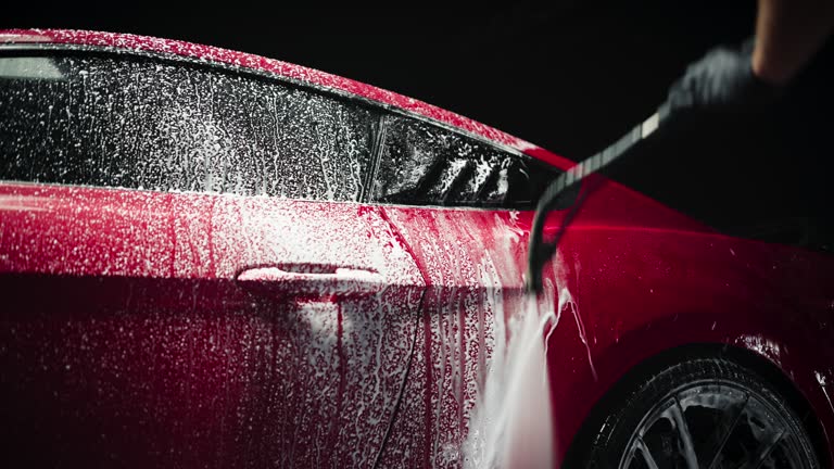 Automotive Detailer Washing Away Smart Soap and Foam with a Water High Pressure Washer. Close Up of a Luxurious Red Supercar Getting Care and Treatment at a Professional Vehicle Body Repair Shop