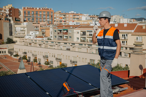Technical engineer in orange vest and helmet tests urban household solar panels with multimeter while talks with Portable radio transceiver. Photovoltaic system installation on the roof of a city.