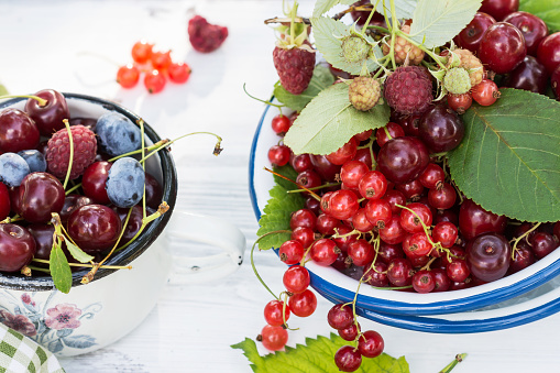 Freshly gathered juicy red currants, cherries, raspberries, blueberries in white plate and cup in garden on sunny day close up, berries on white wooden table background, harvest of berries concept