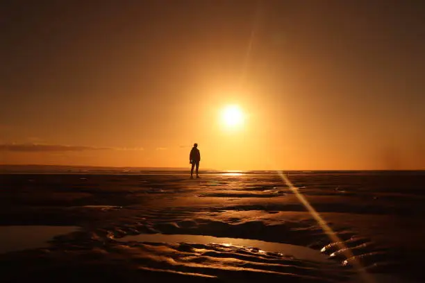 Ambient orange sunset making a silhouette of a man standing on the beach alone with a bright orange sky and waves and sea and coast in background, thoughtful meaningful, epic pose