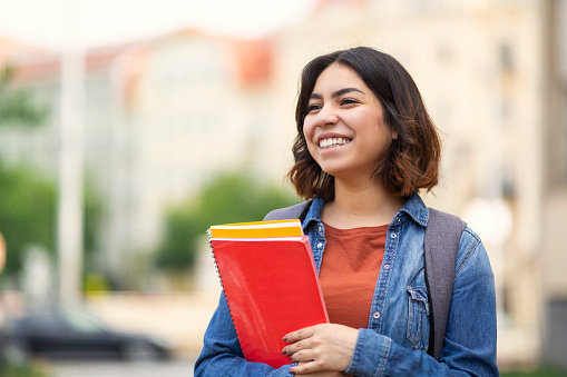 Smiling Middle Eastern Female Student With Workbooks In And Backpack Standing Outdoors, Portrait Of Young Cheerful Arab Woman Posing Outside On City Street, Holding Notepads And Looking Aside