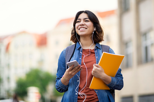 Cheerful young arab student female listening music with smartphone and earphones outdoors, smiling middle eastern woman carrying workbooks and backpack while walking on city street, copy space