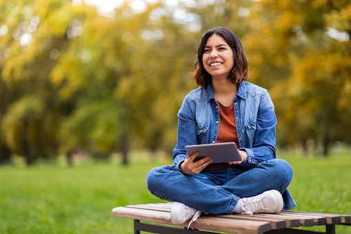 Happy Beautiful Young Arab Woman Relaxing With Digital Tablet Outdoors, Cheerful Middle Eastern Female Sitting on Bench In Park And Using Modern Gadget, Browsing Internet Or Checking Social Media
