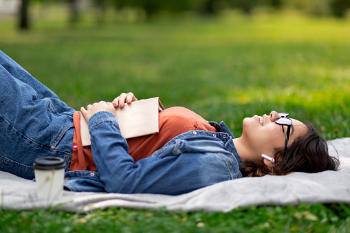 Young Arab Woman Relaxing On Lawn In Park With Book And Earphones, Smiling Middle Eastern Female Lying With Eyes Closed And Listening Music, Daydreaming And Resting On Blanket Outdoors, Side View