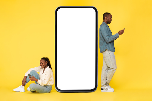 Cheerful Black Couple Texting On Mobile Phones Posing Near Large Smartphone With Empty Screen For Application Advertisement Over Yellow Studio Background. Full Length, Mockup