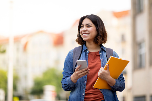 Cheerful Arab Female Student With Smartphone And Workbooks Standing Outdoors, Happy Young Middle Eastern Woman Walking In City After College Classes, Looking Away And Smiling, Copy Space