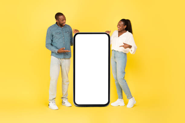 istock Black Spouses Pointing At Large Empty Cellphone Screen, Yellow Background 1473893145