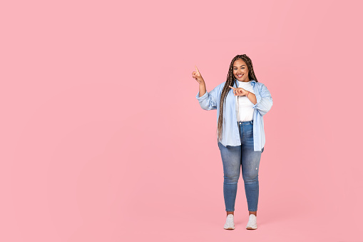 Look There. Full Length Shot Of Happy Black Obese Woman Pointing Fingers Aside At Free Space For Text Standing On Pink Studio Background. Female Advertising Great Offer