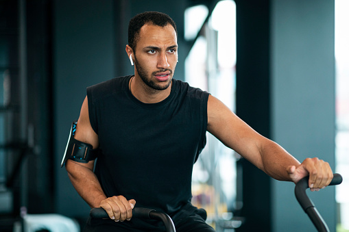 Motivated Black Man Using Elliptical Bike Machine While Training At Gym, Young African American Male Athlete Doing Cardio Workout At Modern Fitness Studio, Enjoying Sporty Lifestyle, Closeup